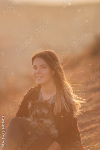 A modern girl walks with Schnauzer through the autumn hills. Sits on the burnt grass, hugs the dog with love, care in the sunset, smiles, looks at the camera, paragliders fly behind her. Traveling pet