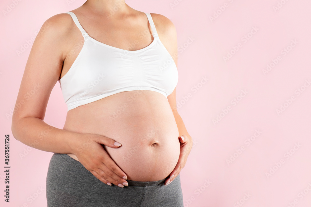 Close up of pregnant woman wearing supportive seamless maternity bra & grey yoga pants, arms on her belly. Female hands wrapped around big bare tummy. Child expectancy concept. Background, copy space Close up of pregnant woman wearing supportive seamless maternity bra & grey yoga pants, arms on her belly. Female hands wrapped around big bare tummy. Child expectancy concept. Background, copy space