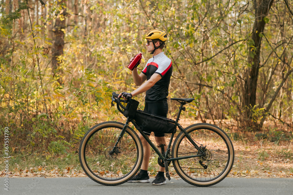Man in sportswear standing in autumn forest with bicycle and bottle of water in his hands and looking away. Athlete weightlifter is resting in training, drinking water from a bottle.