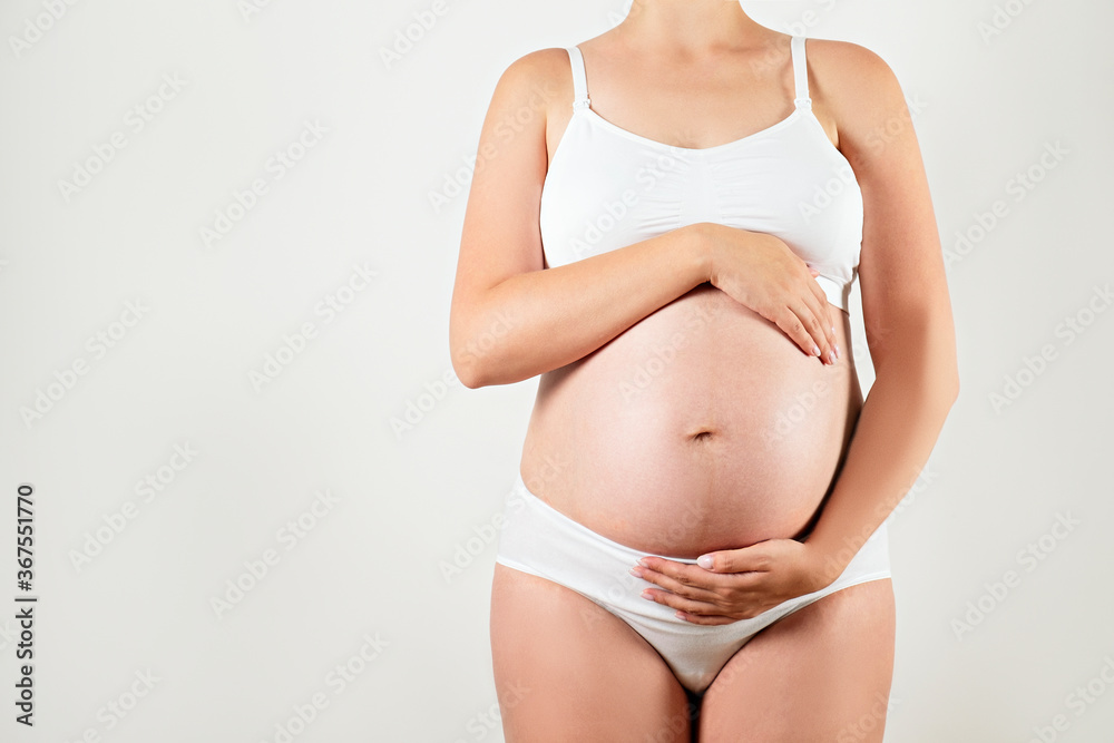 Close up of pregnant woman wearing supportive seamless maternity bra and maxi bottoms, arms on her belly. Female hands wrapped around big bare tummy. Child expectancy concept. Background, copy space Close up of pregnant woman wearing supportive seamless maternity bra and maxi bottoms, arms on her belly. Female hands wrapped around big bare tummy. Child expectancy concept. Background, copy space