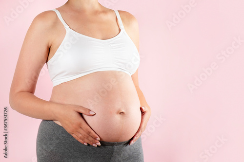 Close up of pregnant woman wearing supportive seamless maternity bra & grey yoga pants, arms on her belly. Female hands wrapped around big bare tummy. Child expectancy concept. Background, copy space © Evrymmnt