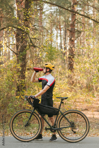 Fototapeta Naklejka Na Ścianę i Meble -  Male cyclist in a sports outfit walks through an autumn park and quenches his thirst, drinks water from a bottle and looks away. The cyclist is resting in training, drinking water. Vertical