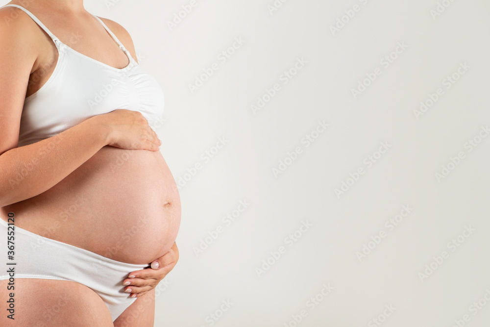 Close up of pregnant woman wearing supportive seamless maternity bra and maxi bottoms, arms on her belly. Female hands wrapped around big bare tummy. Child expectancy concept. Background, copy space Close up of pregnant woman wearing supportive seamless maternity bra and maxi bottoms, arms on her belly. Female hands wrapped around big bare tummy. Child expectancy concept. Background, copy space