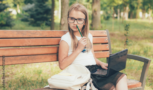 Schoolgirl in glasses sits on a bench in the park with a laptop and learns remotely