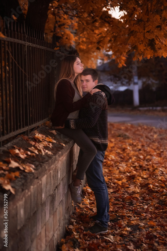 A young couple of teenagers are sitting by the fence under the orange foliage of a tree in the autumn evening. Lovers hug, look at each other, kiss. First date, weekend travel together. Walking night