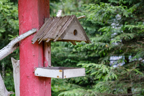 A two-piece wooden bird feeder nailed to a red-painted post, with dense trees in the background.