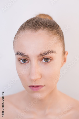 Close-up portrait of a blonde girl with nude makeup, hair gathered in a bun, naked against a gray wall. Natural beauty concept without surgery. Clean face, well-groomed skin, healthy appearance. 