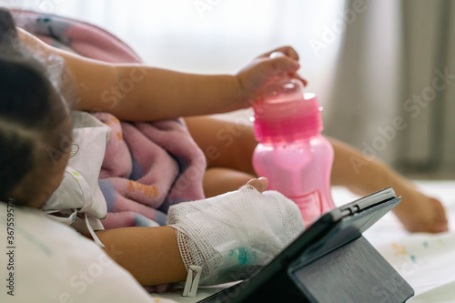 Child or girl patient rest on bed with milk bottle and play or watch smart tablet while hand still lock with equipment medical for injection for saline or vitamin or infusion intravenous