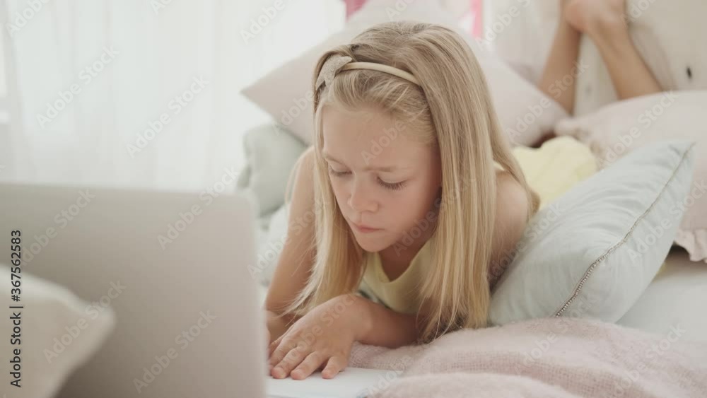 Blonde girl 10 years old is doing homework in bed in front of a laptop