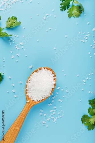 Sea salt in a wooden spoon on a blue background, with salt and herbs scattered around the edges.