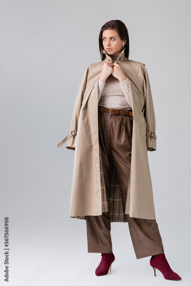 full length view of fashionable girl touching trench coat and looking away on grey full length view of fashionable girl touching trench coat and looking away on grey