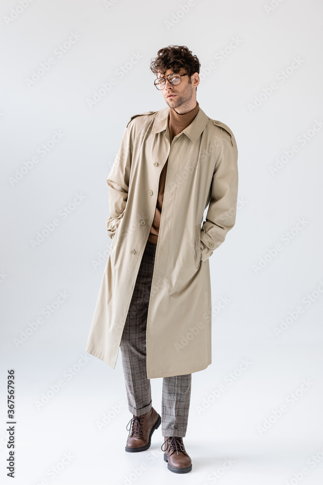 full length view of fashionable man in trench coat holding hands in pockets  while posing on grey Photos | Adobe Stock