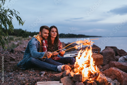 happy couple roasting marshmallows on sticks near bonfire