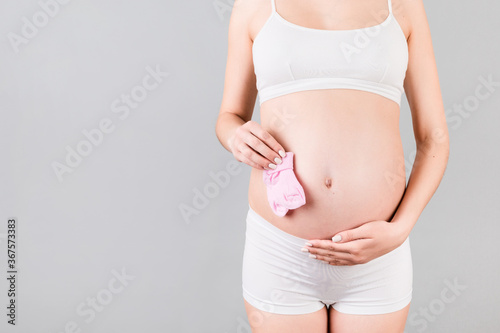 Cropped image of pink socks for a baby girl in pregnant woman's hands against her belly at gray background. Parenthood concept. Copy space
