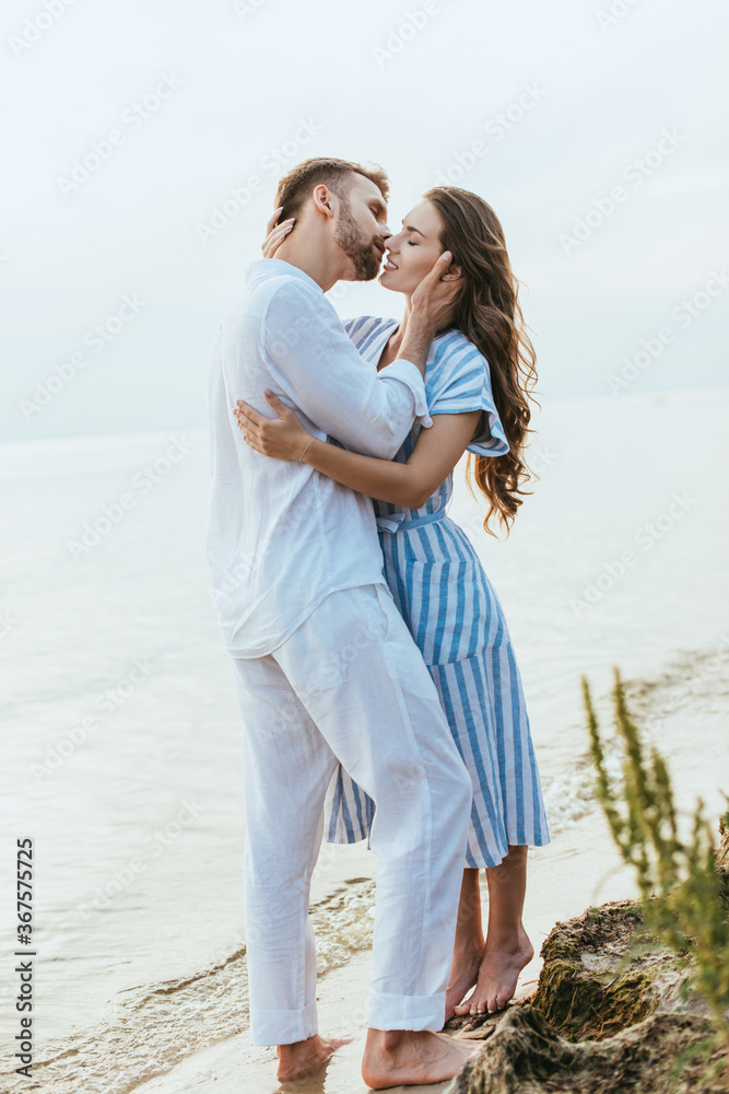 selective focus of happy man hugging and kissing girl in dress near lake selective focus of happy man hugging and kissing girl in dress near lake