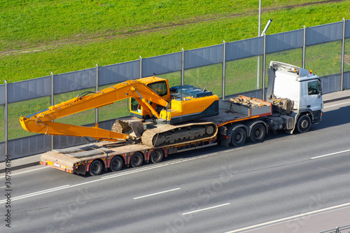 Heavy new yellow excavator long boom bucket on transportation truck with long trailer platform on the highway in the city.