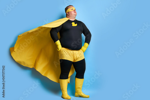 handsome brave strong man has a mad desire, a dream to shield the world from dirt and evil. fat superhero in yellow costume posing isolated over blue background