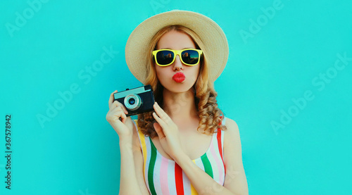 Portrait of young woman photographer with vintage film camera blowing red lips sending sweet air kiss wearing a summer straw hat over blue background