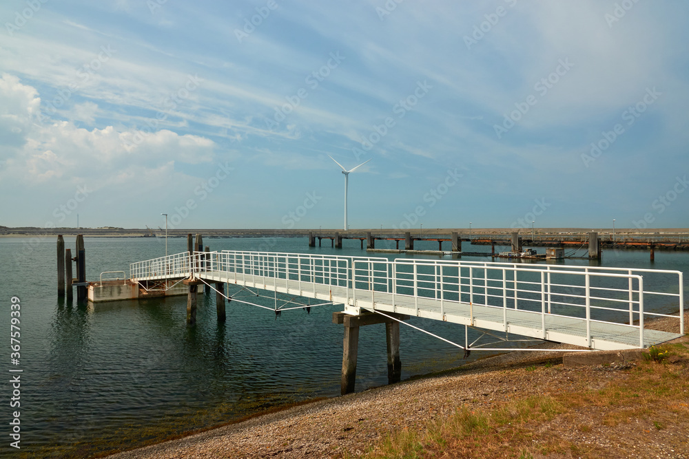Long white pier leads to the boat dock. Wind turbine and dam in the background. Zeeland, Netherlands.