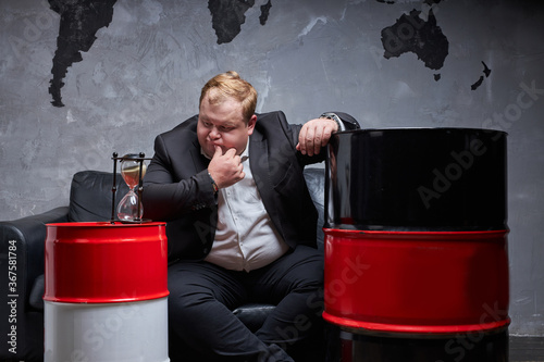 fat caucasian oil businessman thinks how to solve his oil business, he has no time. oil barrels next to him. oil industry, crisis concept photo