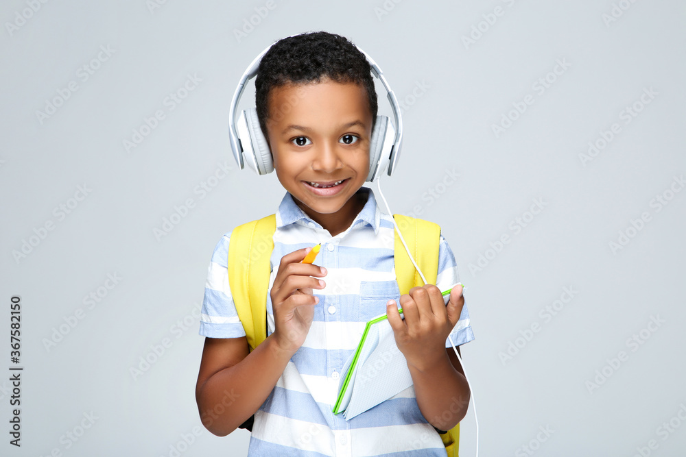 Young African American school boy writing in notepad with headphones and backpack on grey background Young African American school boy writing in notepad with headphones and backpack on grey background