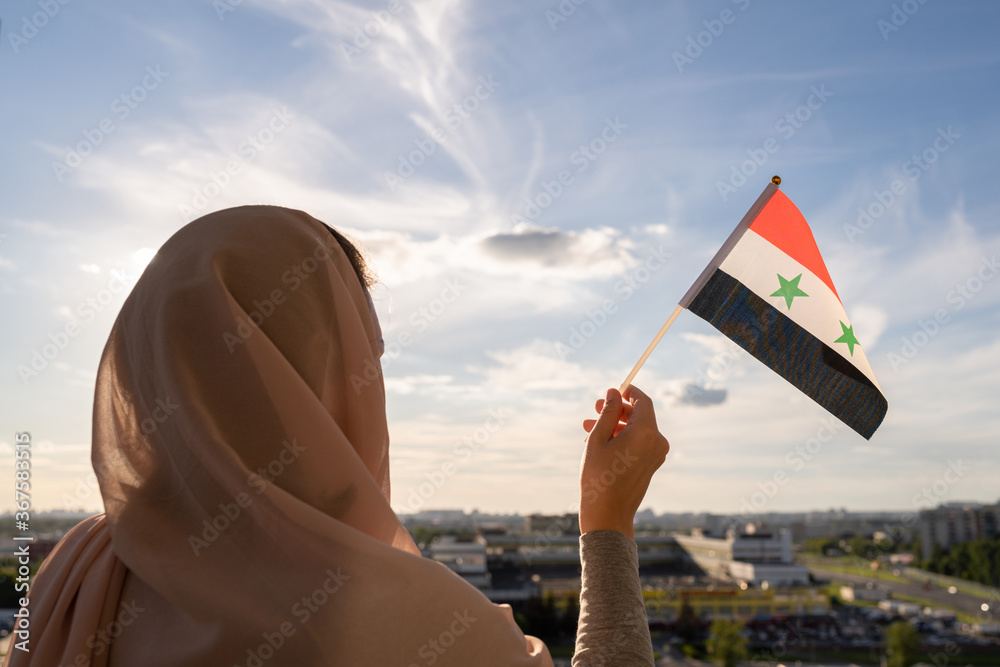 Fototapeta premium Silhouette of muslim woman in head scarf with Syria flag at blue sunset sky. Concept of freedom