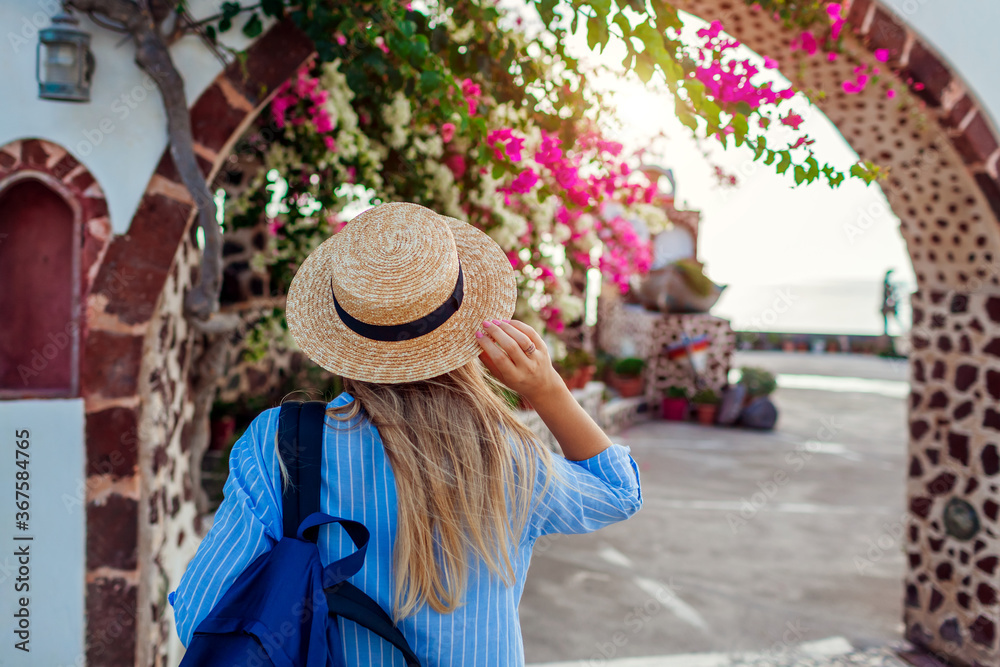 Woman traveler walks along traditional village architecture on Santorini island. Tourist admires flowers. Summer