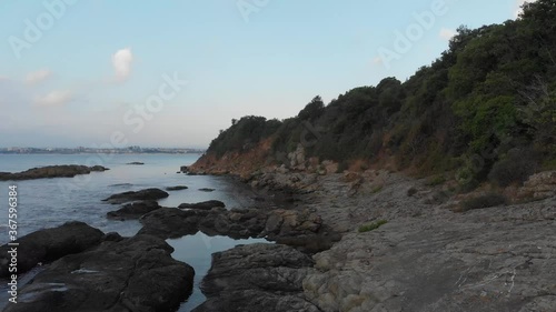 Rocky coastline of the sea at the foot of a steep high cliff, which ends with a dense forest.