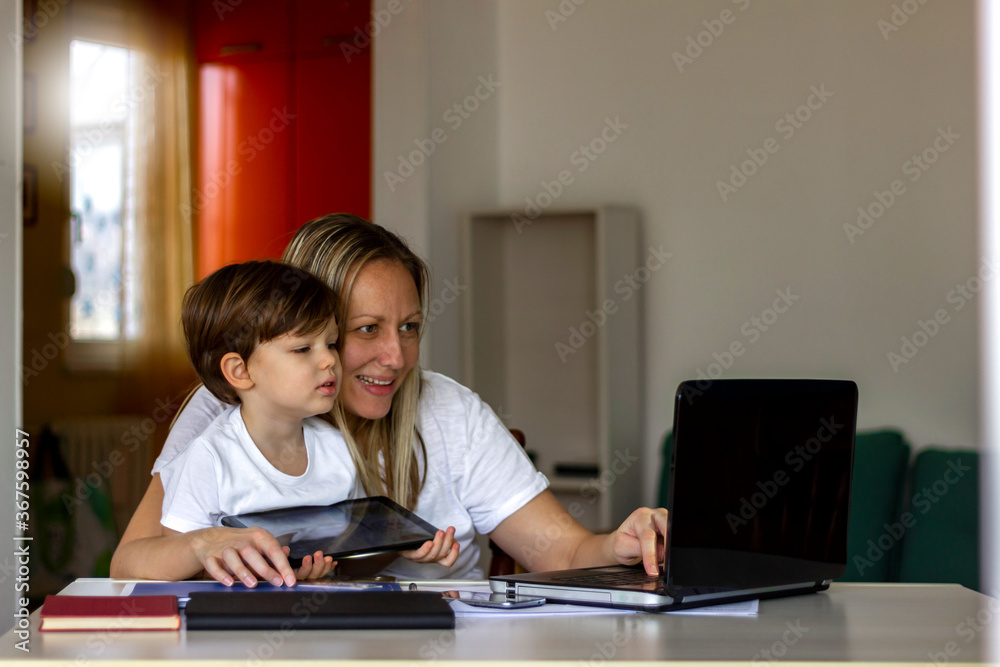 Young Caucasian single mother working on laptop while her son is using digital tablet.Mother and her son spending time together at home.Mother using laptop and her son using tablet.Technology concept. Young Caucasian single mother working on laptop while her son is using digital tablet.Mother and her son spending time together at home.Mother using laptop and her son using tablet.Technology concept.