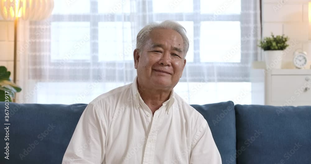 Closeup portrait of healthy cheerful asian senior elderly old gray-haired man smiling to the camera while sitting on the blue couch in the living room. happy mature middle aged grandfather. Closeup portrait of healthy cheerful asian senior elderly old gray-haired man smiling to the camera while sitting on the blue couch in the living room. happy mature middle aged grandfather.