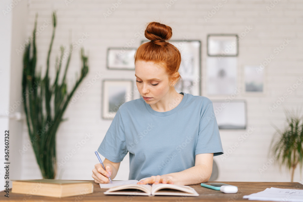 Focused redhead young woman is writing letter on table at home office. Concentrated businesswoman making notes in notebook at the desk in light cozy living room. Focused redhead young woman is writing letter on table at home office. Concentrated businesswoman making notes in notebook at the desk in light cozy living room.