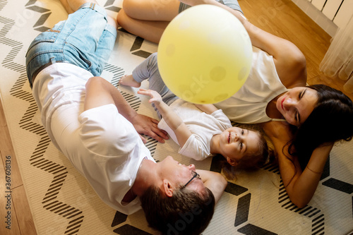 Pregnant mom, dad and daughter 2 years old play ball, hug and rejoice lying on the carpet in the living room at home