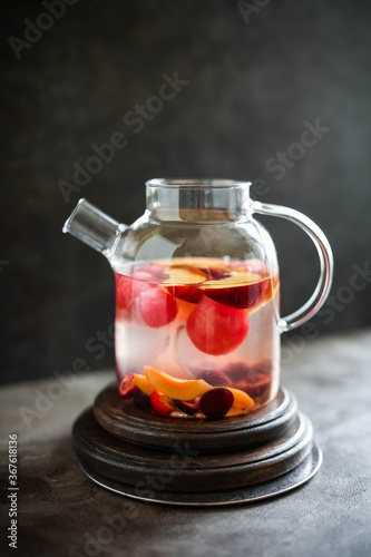 Fruit tea in a large transparent teapot. Warm lemonade with fruits and berries. Dark photo. Minimalism. Beautiful dishes.