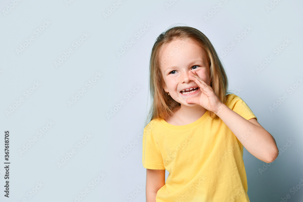 Cute little girl in bright yellow t-shirt holding hand close to mouth sharing rumor or secret. Waist up shot isolated on light blue, copy space Cute little girl in bright yellow t-shirt holding hand close to mouth sharing rumor or secret. Waist up shot isolated on light blue, copy space