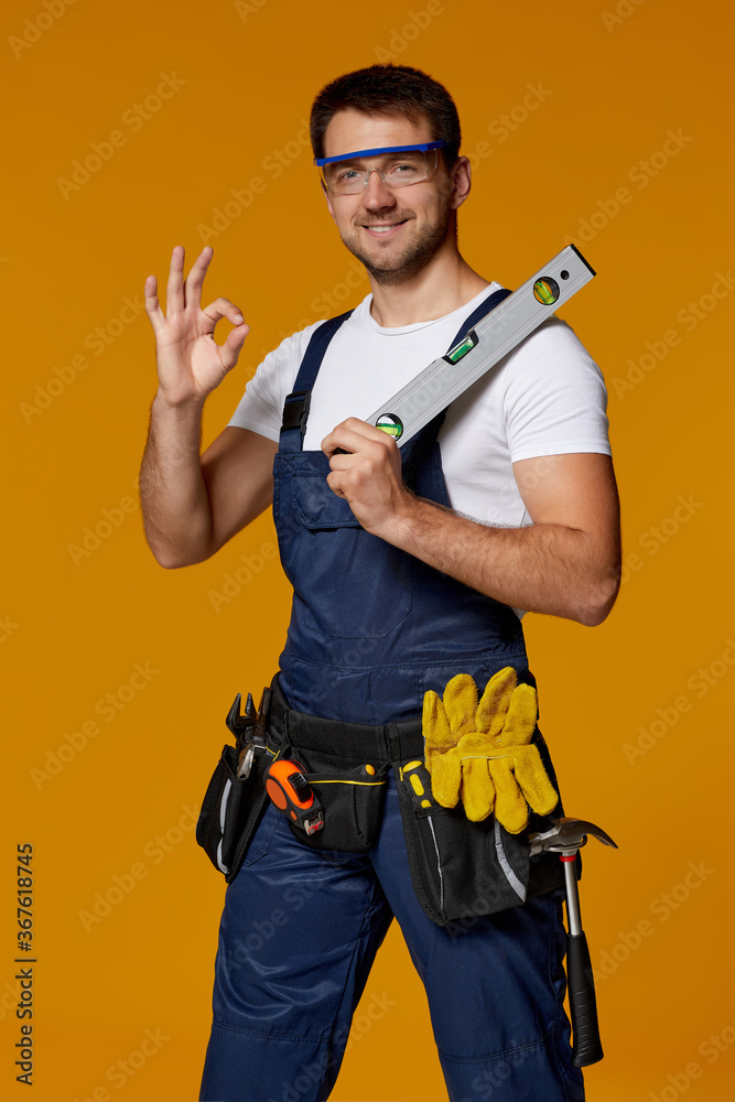 Young caucasian repairman worker in safety glasses and uniform holding builder level. handyman wearing tool belt showing Ok gesture isolated on orange studio background - obrazy, fototapety, plakaty 