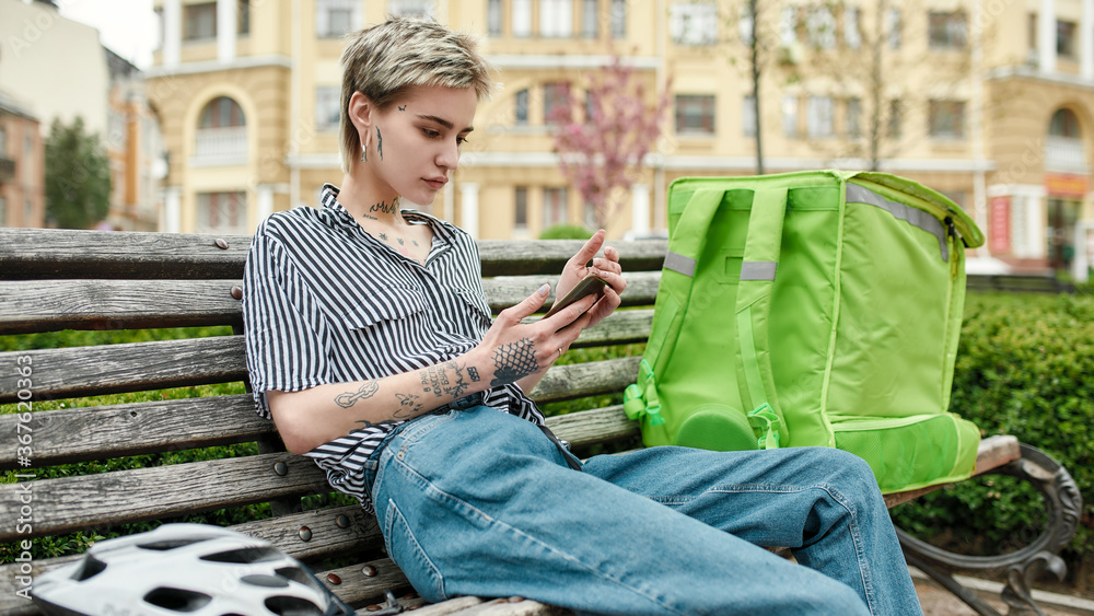 Obraz premium Young delivery woman with thermo bag or backpack checking order using smartphone, sitting on the bench outdoors. Courier, delivery service concept