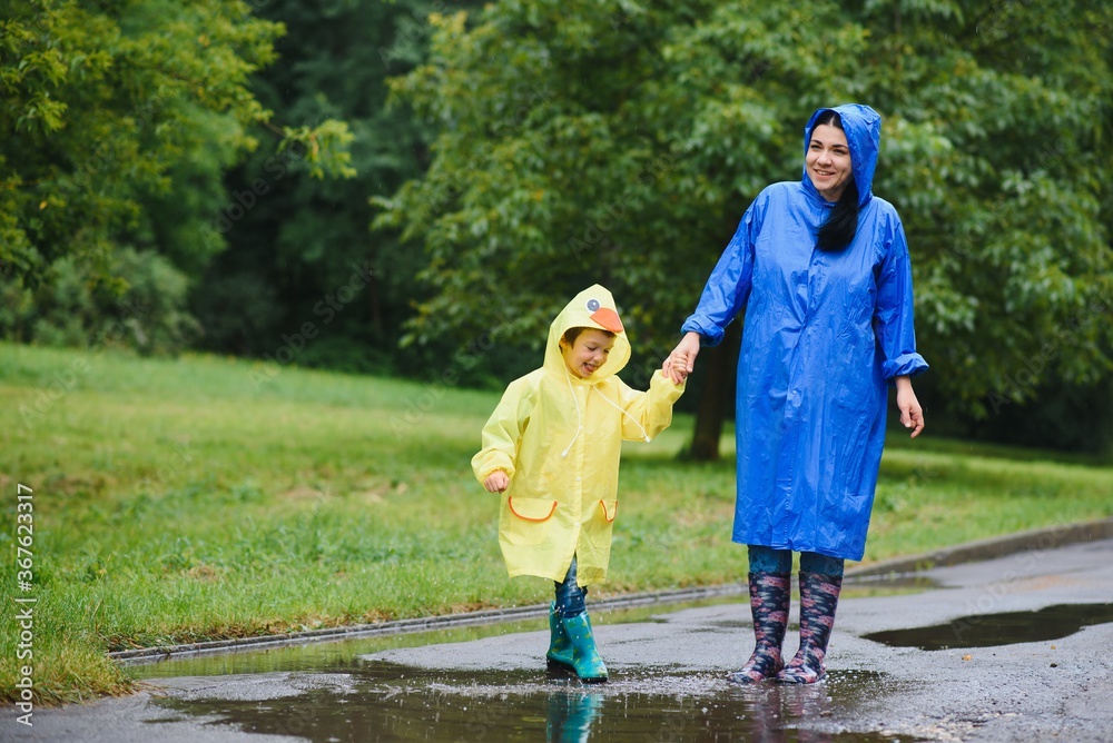 Mother with son walking in park in the rain wearing rubber boots - obrazy, fototapety, plakaty 