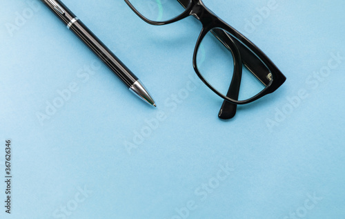 Glasses and a pen are on the table. Close up top view of pen and glasses on blue background for mockup design modern lifestyle stationary concept. Blue office desk with supplies. Space for your text.