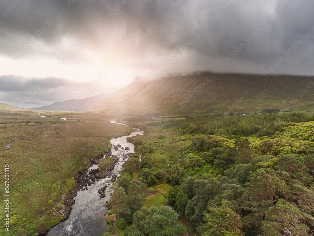 Aerial drone view on a landscape in Connemara region. county Galway, Ireland, Small river flows from mountain by a forest, Sun shines over mountain, Cloudy sky.