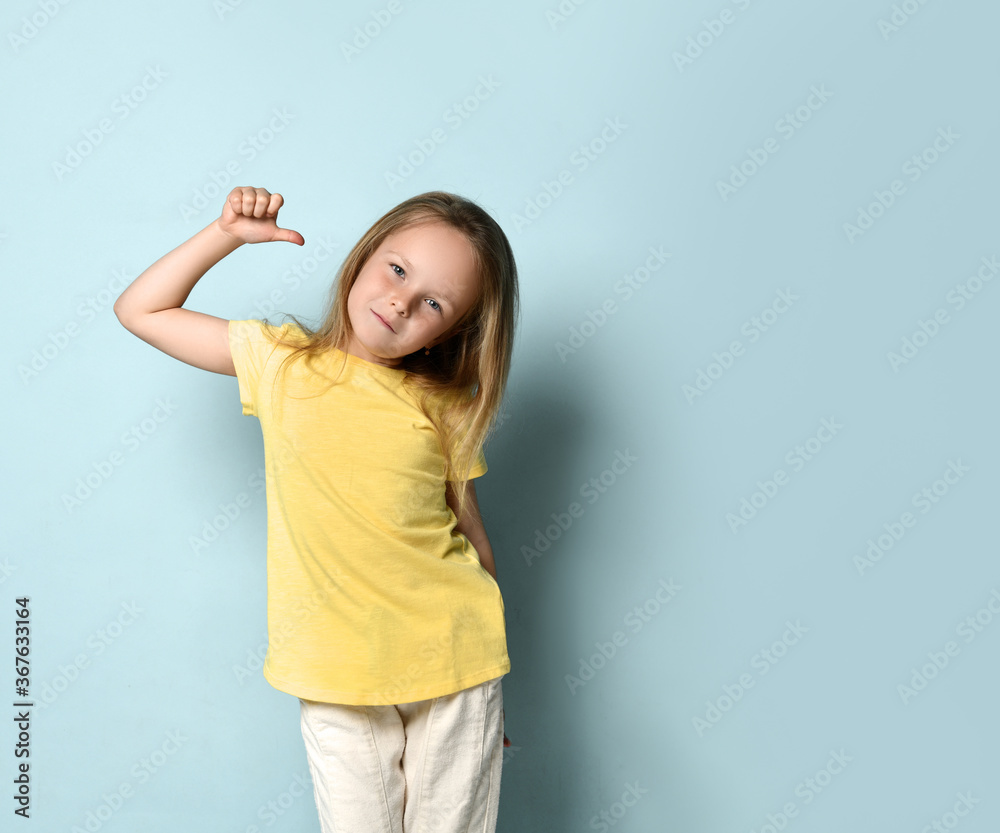 Confident little preschool girl posing in yellow t-shirt pointing with thumbs at herself. Waist up shot isolated on light blue Confident little preschool girl posing in yellow t-shirt pointing with thumbs at herself. Waist up shot isolated on light blue