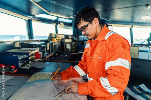 Filipino deck Officer on bridge of vessel or ship wearing coverall during navigaton watch at sea . He is plotting position on chart