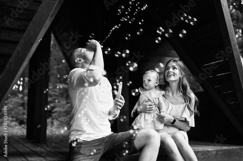 Happy family play outdoor, caring father blow the bubbles for baby girl to laugh, overjoyed mom and daughter looking at the bubbles, smiling, funny parenting moments concept