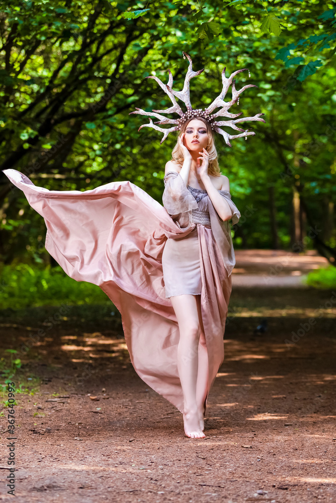 Fototapeta premium Young Beautiful Girl Posing With Artistic Deer Horns In Summer Forest and Wearing Flying Light Dress as Forest Nymph Concept. Art Photography.