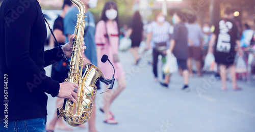 A street musician plays the saxophone and wearing face shield with blurry many people wearing mask and walking in Bangkok, Thailand.