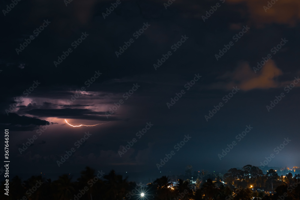 Thunderstorm, lightning and orange clouds in the dramatic night sky over the sea, trees and houses on a tropical island