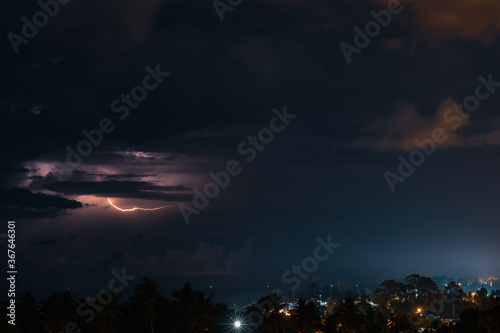 Thunderstorm  lightning and orange clouds in the dramatic night sky over the sea  trees and houses on a tropical island