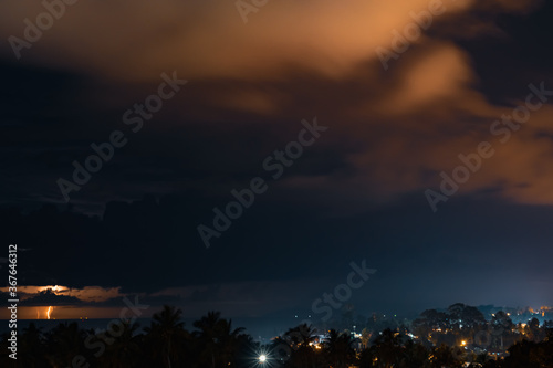 Thunderstorm, lightning and orange clouds in the dramatic night sky over the sea, trees and houses on a tropical island