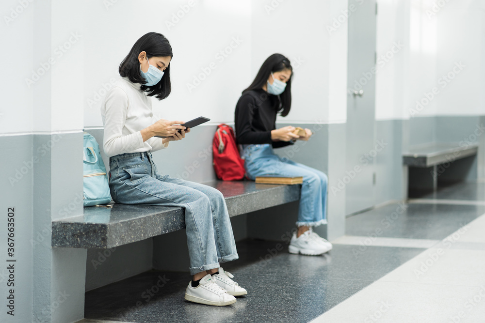 Two female college students wearing mask, reading book and learning while keeping social distance in university campus during COVID-19 pandemic. Stock photo.