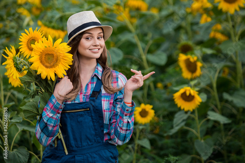 Gorgeous, adorable, energetic, female farmer pointing to her left while holding many sunflowers, in the middle of a green and yellow sunflower field.