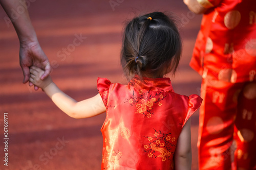 Asian sister walk and hold mom hand behind brother in traditional red cheongsam costume walking together in park. 2021 Chinese new year holiday concept. Happy China family.